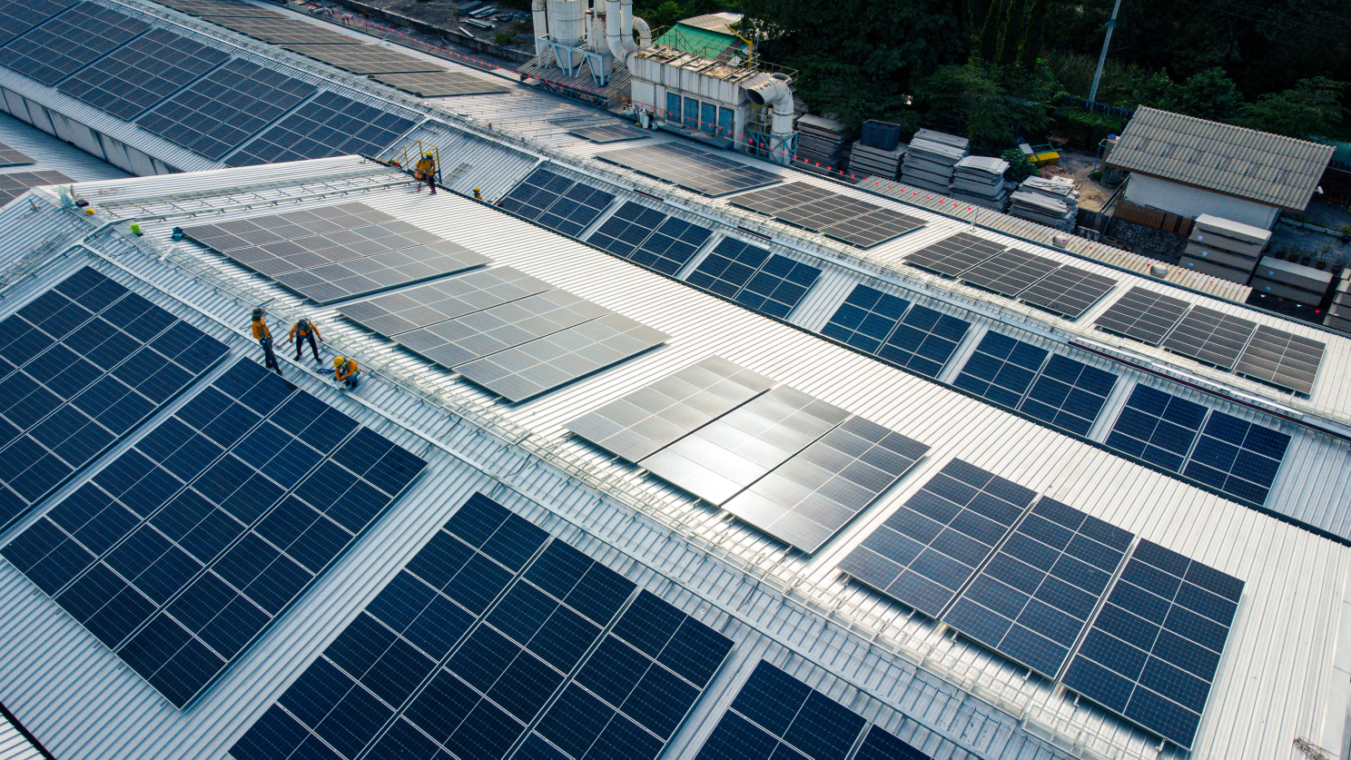 Aerial view of workers installing rooftop solar panels on a large industrial facility, showing distributed renewable assets that feed into Virtual Power Plants for ancillary services revenue stacking
